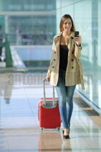 Front view of a traveler woman walking and using a smart phone in an airport corridor