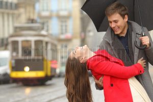 Happy playful couple in love dating joking and laughing in the street of an old town in a rainy day under an umbrella